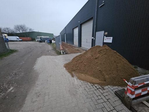 Een hopen zand naast een gebouw op een grindpad. Achtergrond met voertuigen en een hek.